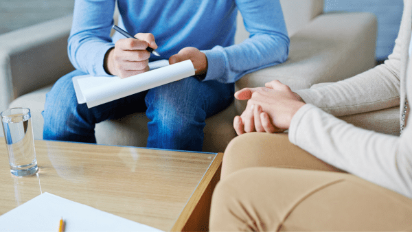 counseling session between 2 people. One is writing while the other has hands in lap